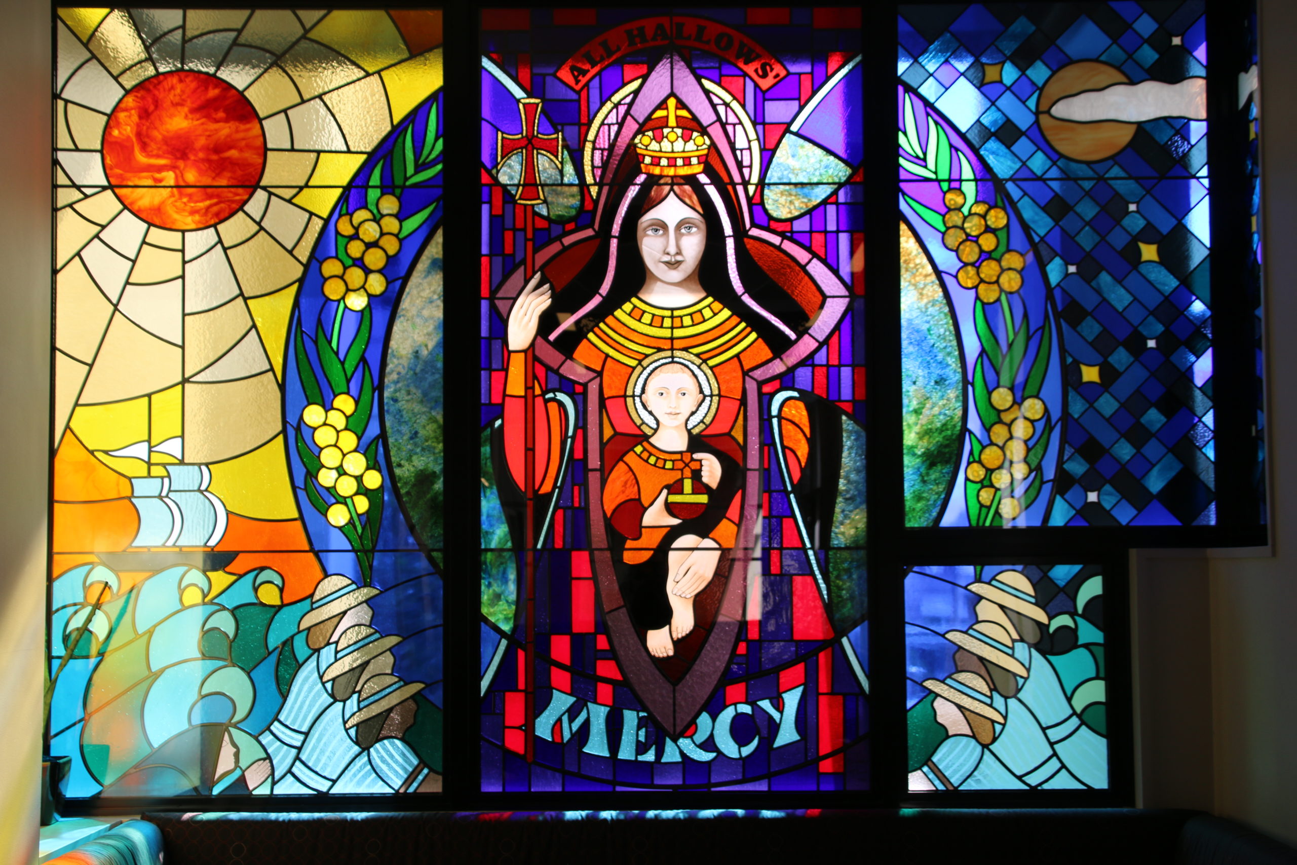 This is a photo of a beautiful coloured stained glass window from All Hallows' School. At the centre of the window is the Virgin Mary with baby Jesus sitting on her lap. The design also includes wattle branches, a shining sun, a boat and waves, and All Hallows' school students.