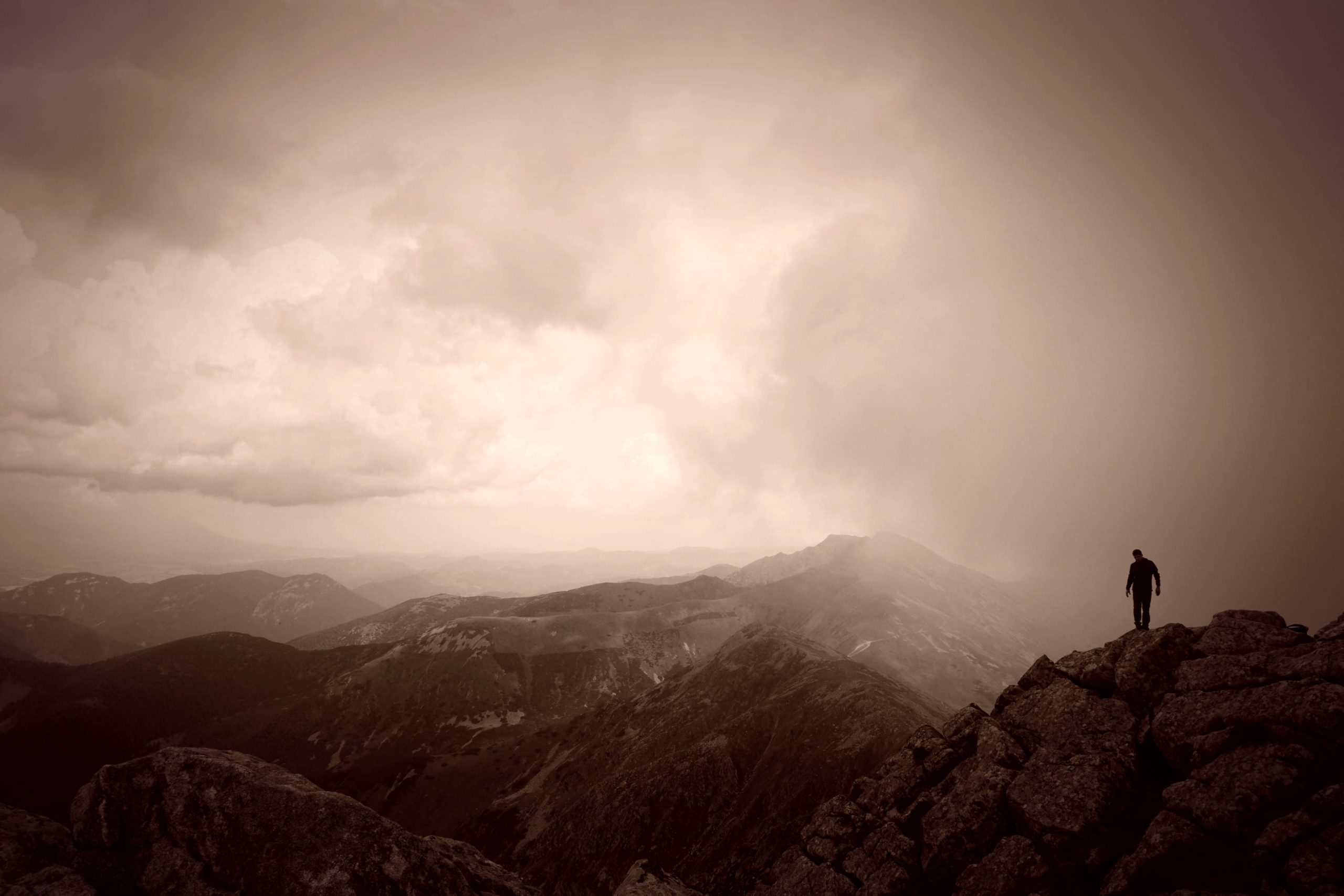 Sepia toned photograph of a man standing on top of a cloud covered mountain looking out over a mountain range
