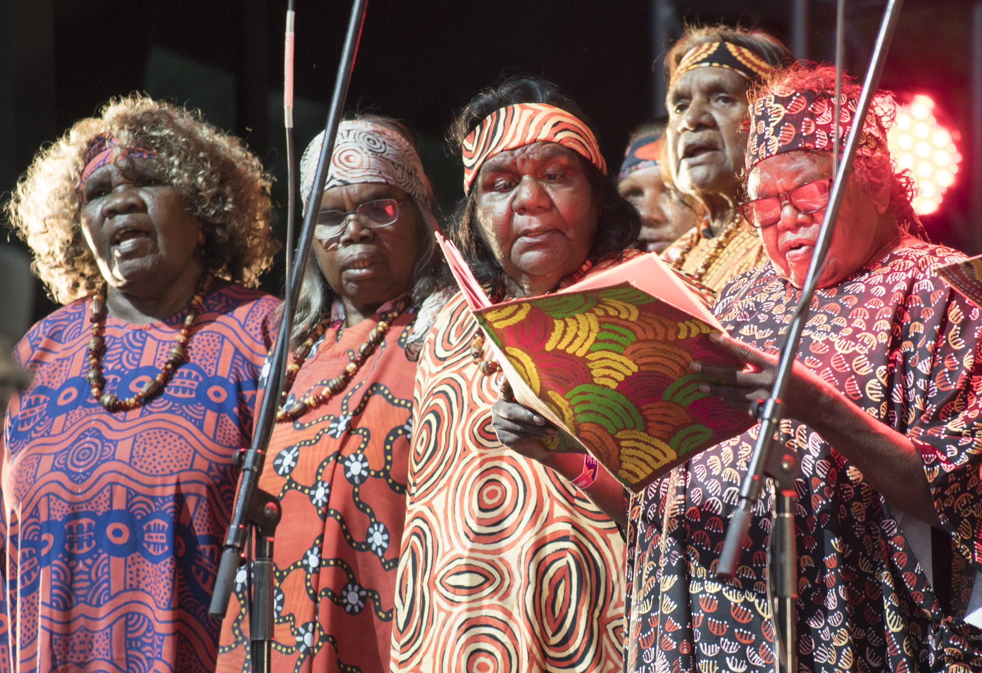Aboriginal elder women singing in a choir wearing colourful aboriginal designed dresses with matching headbands.