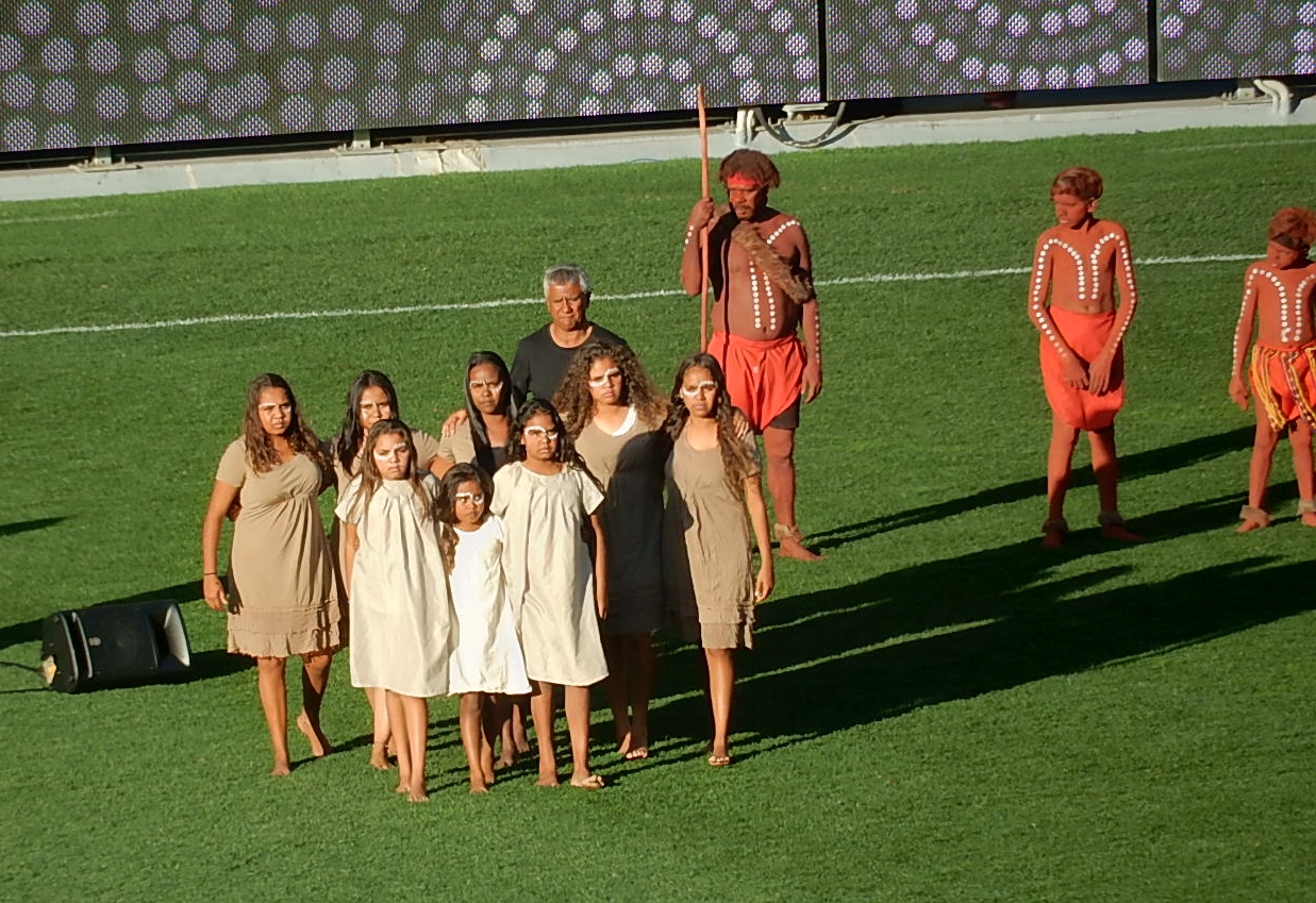 Female Aboriginal youth all dress in variations of beige and brown, with white paint on their faces. Two aboriginal men stands behind them one in a black t-shirt, the other in traditional dress holding, a spear.