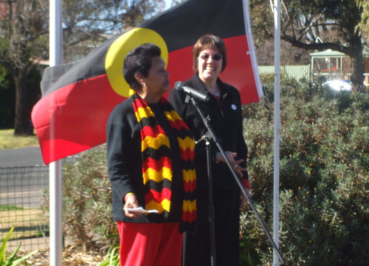 An Aboriginal woman is speaking into a microphone. The Aboriginal flag is flying behind her. She is wearing a scarf in the colours of the Aboriginal flag.