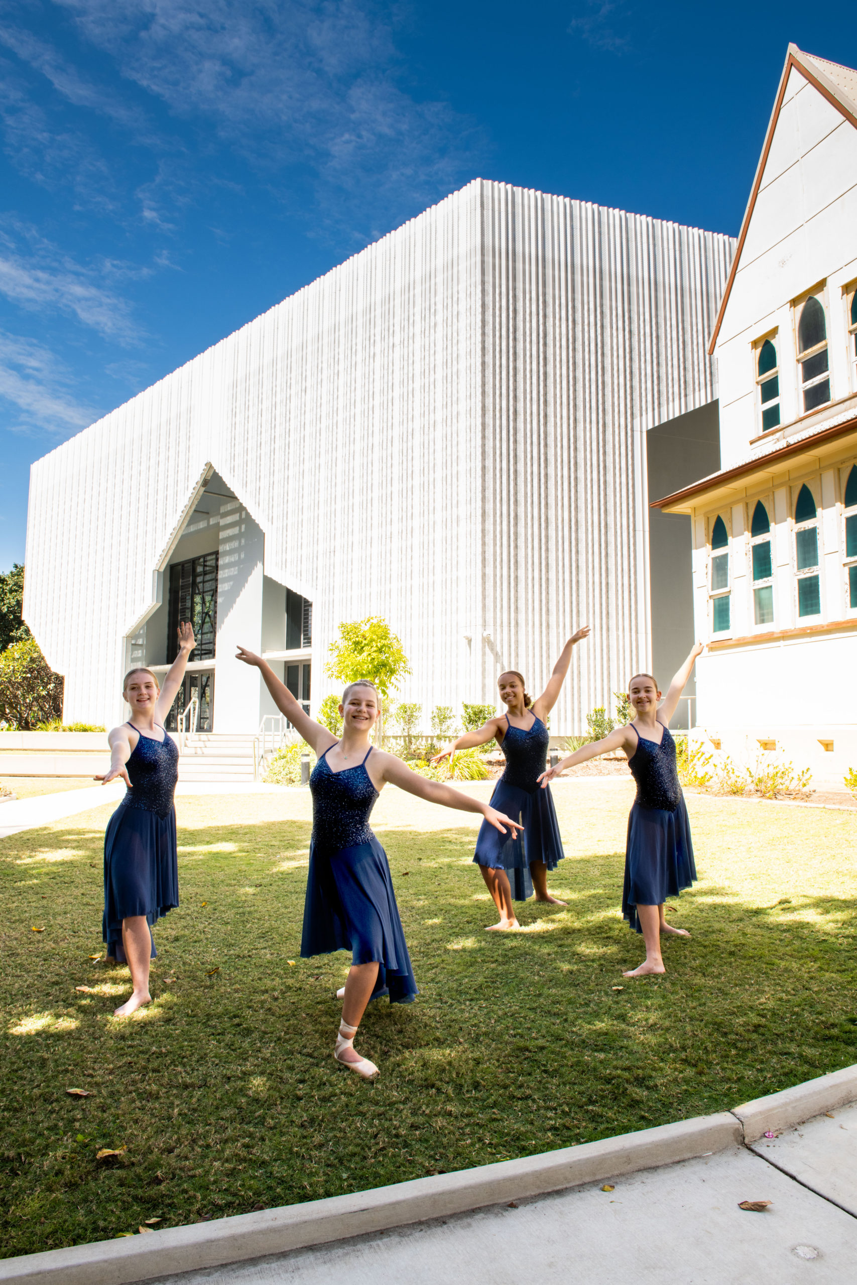 Four dance students from St Patrick's College. They are dressed in blue flowing dresses and wearing ballet shoes and are holding a dance pose on they are on the grass in front of the school.