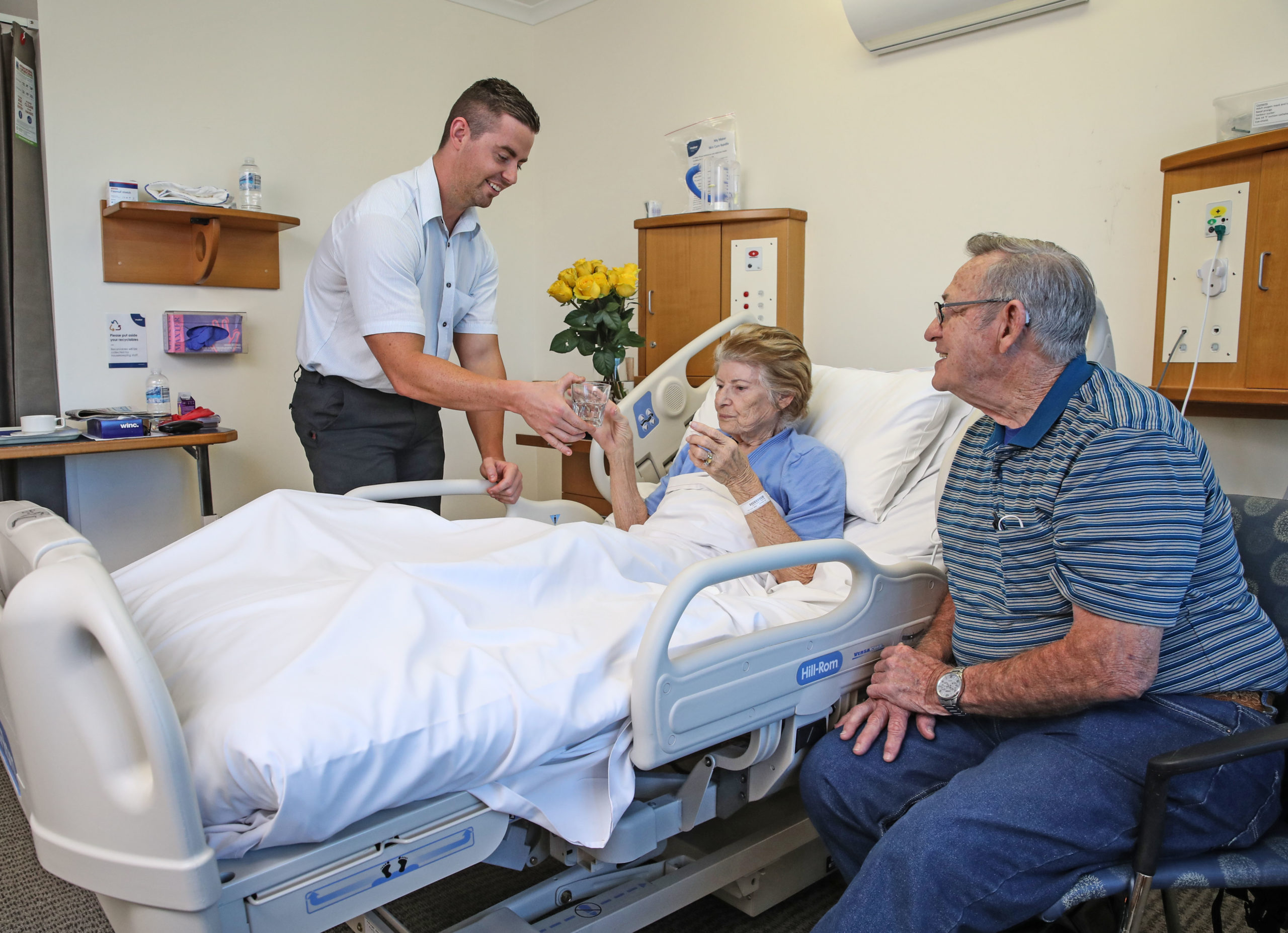 Mater staff caring for an elderly woman. The elderly woman has a visitor with her and elderly man who sits beside her bed.