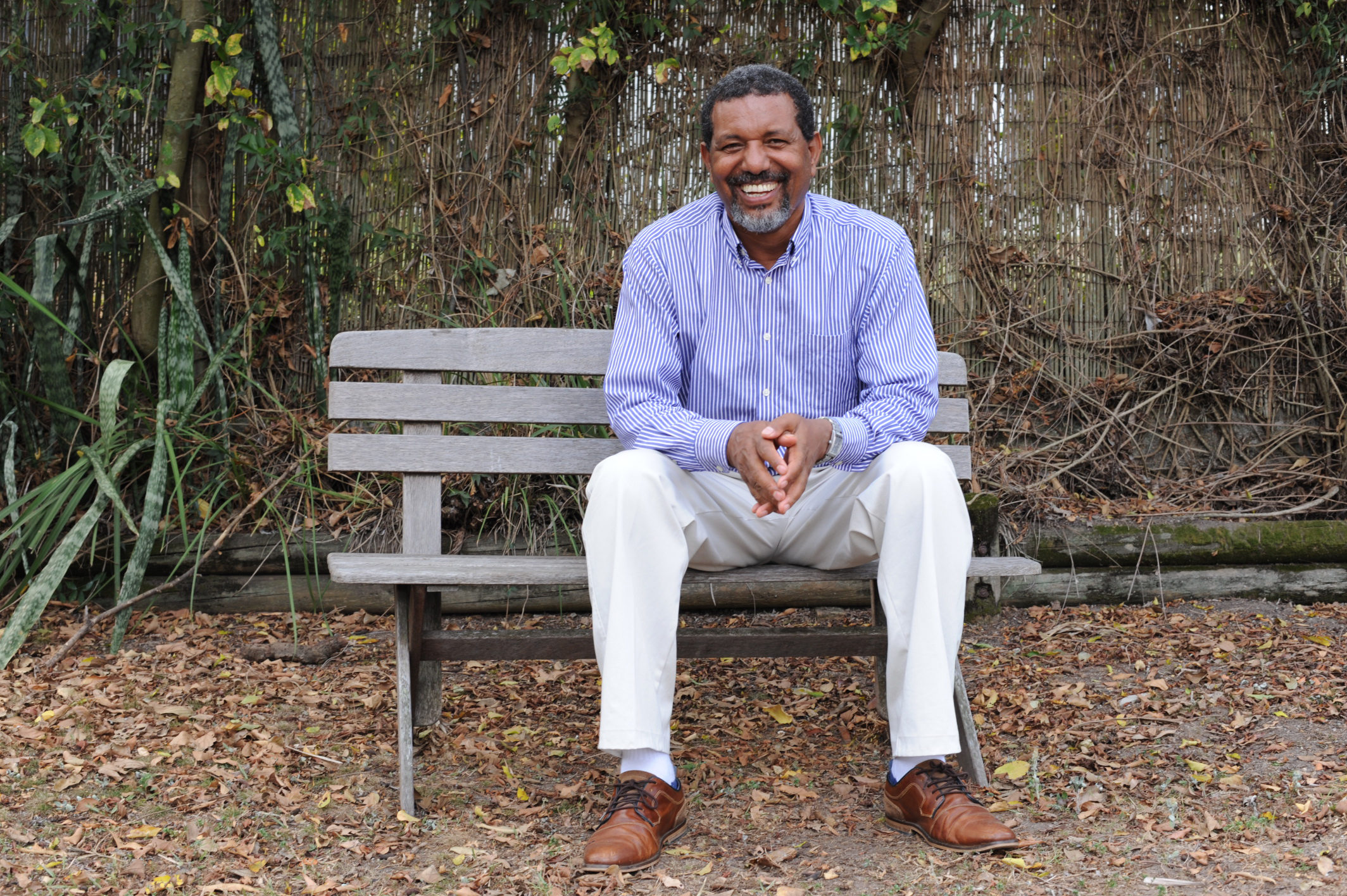 A smiling man sittting on a park bench in autumn, there are browning leaves covering the ground.