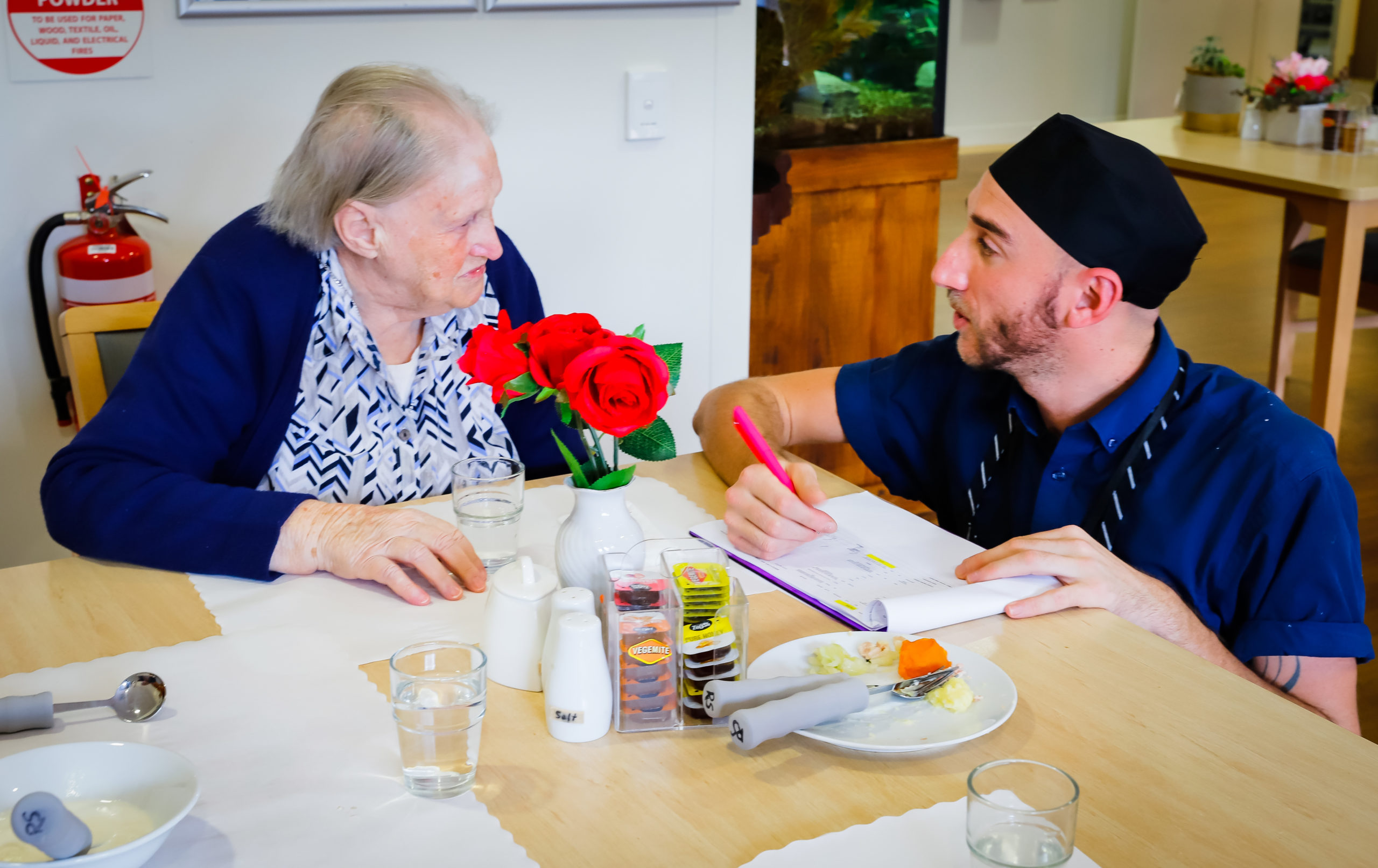 An elderly lady and a young man working talking at a table. The dining table they are at has a white vase with beautiful red roses in it.