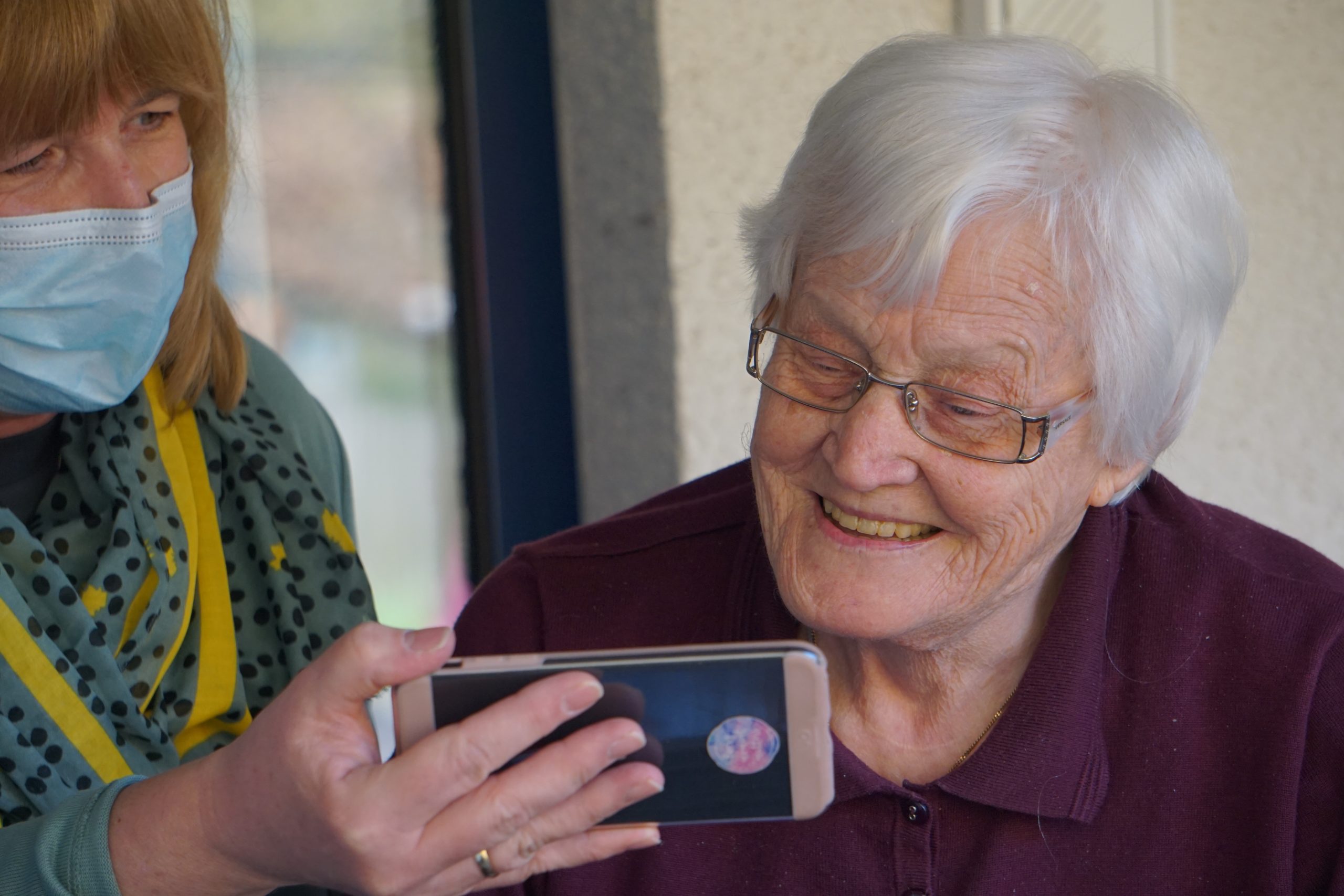 A photograph of a health care worker in a mask assisting an elderly resident to communicate via a smart phone.