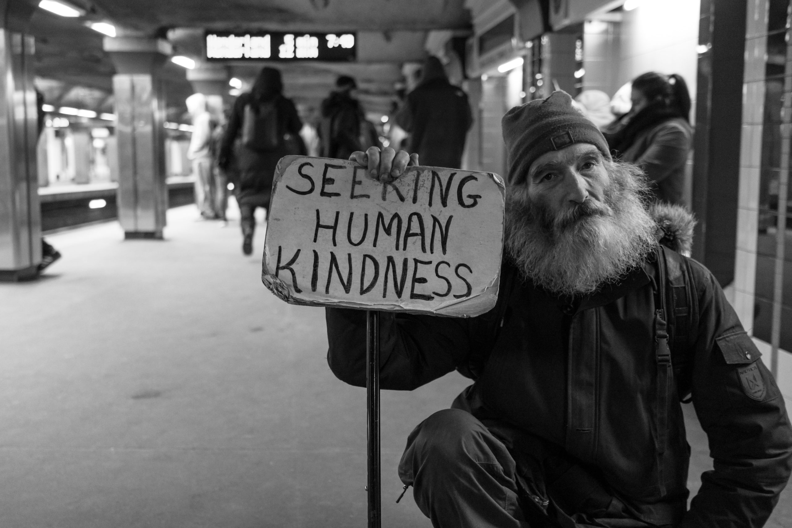 A black and white image of an elderly homeless man holding a sign that reads