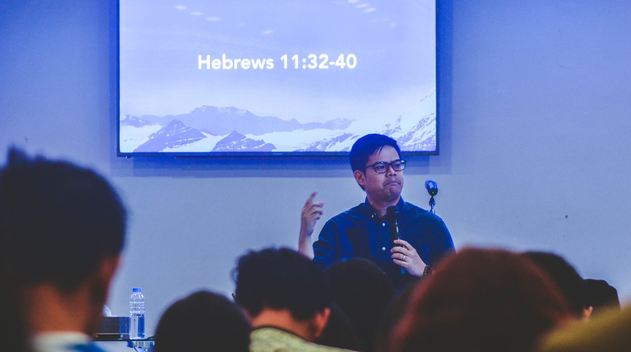 A photograph of a man presenting at a conference. A Bible reference is showing on the screen behind him.