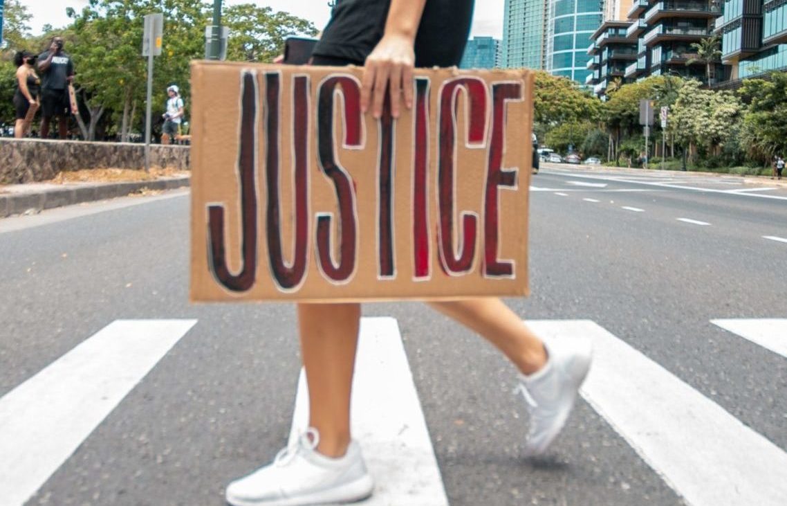 A person walks across a zebra crossing holding a cardboard sign with the word